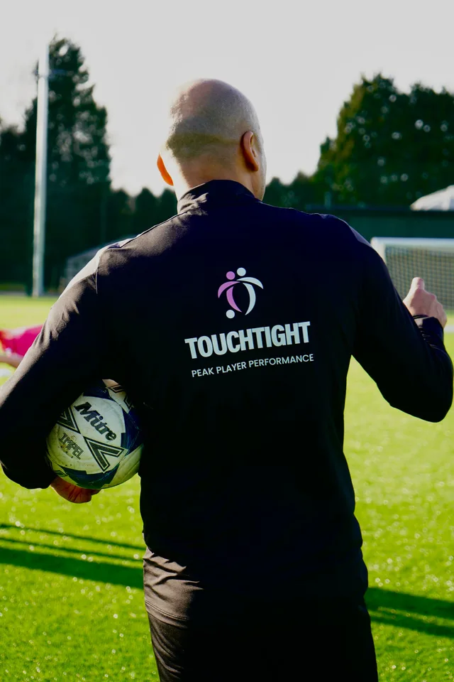 A TouchTight coach seen from behind, holding a ball under floodlights in golden evening light